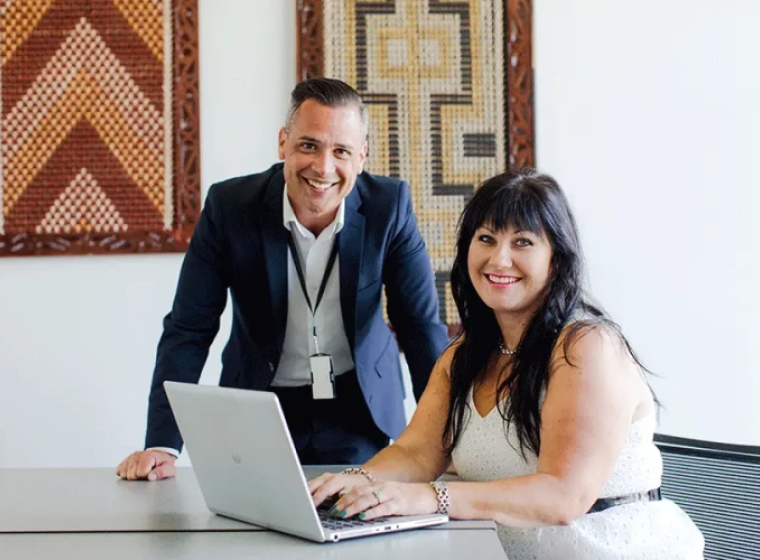 Two health professionals, one leaning on a table, one typing on a laptop